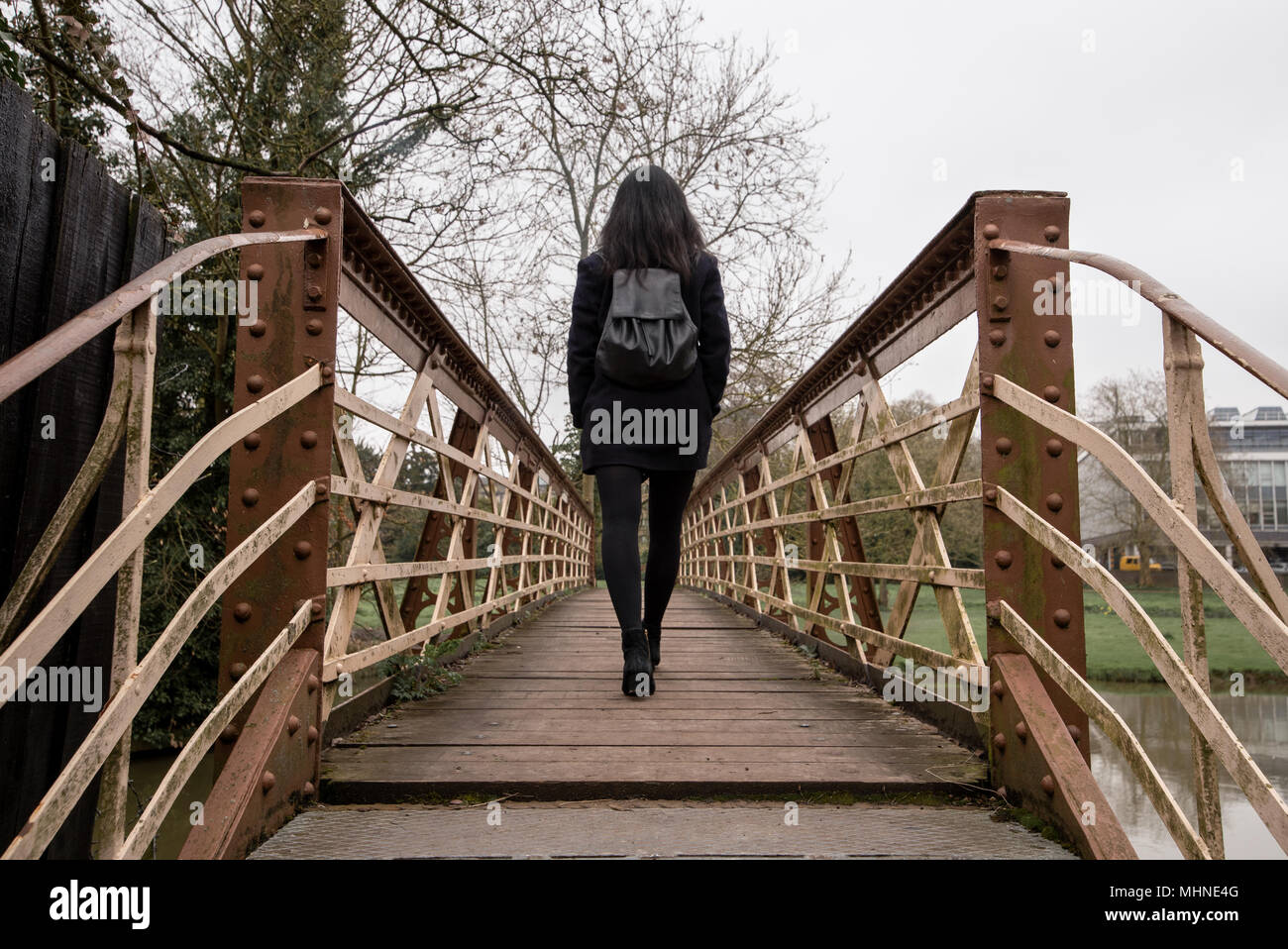 Una giovane donna con i capelli neri e vestito di nero da soli passeggiate attraverso un ponte pedonale in acciaio attraversando un piccolo fiume. Foto Stock