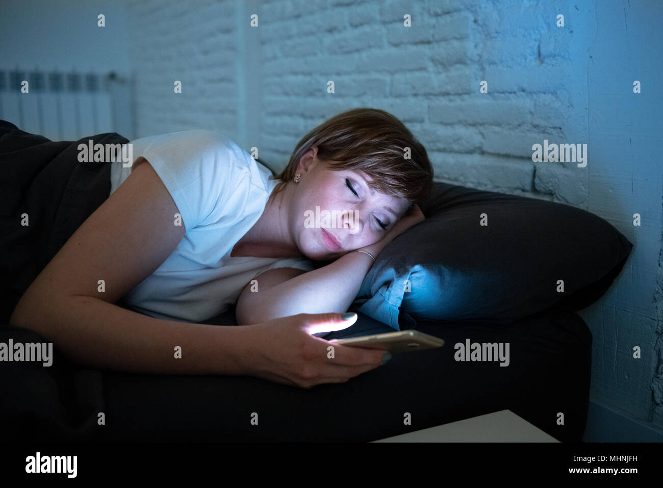 Attraente giovane donna caucasica addormentarsi utilizzando smart phone giacente a letto a tarda notte in una camera oscura. Uso di mobile in dipendenza da internet, mo Foto Stock