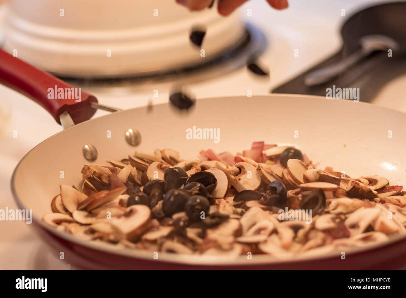 Mano far cadere le olive nere in padella. Un trito di olive nere affettate i funghi e la cipolla rossa in rosso e bianco padella. Foto Stock