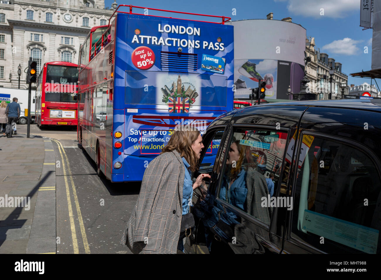 Una donna parla ad un conducente di taxi di fronte a London tour bus con una illustrazione di attrazioni turistiche, con una scheda iVenture salva i visitatori fino al 50% sui biglietti presso la capitale di punti di riferimento, 1 maggio, a Piccadilly Circus a Londra, Inghilterra. Foto Stock