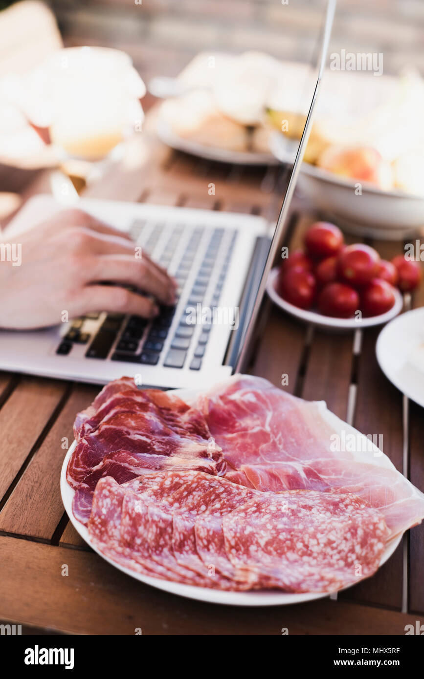 Primo piano di un giovane uomo caucasico usando un computer portatile seduti ad un tavolo di legno all'aperto durante un pasto Foto Stock
