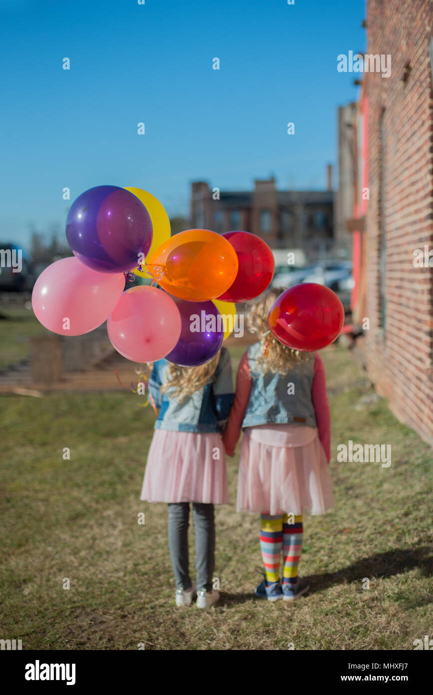 Due sorelle tenendo le mani con grappolo di palloncini colorati, vista posteriore Foto Stock