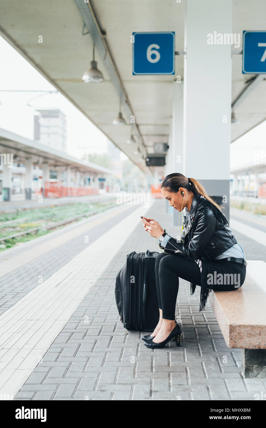 Donna seduta sulla piattaforma del treno, utilizza lo smartphone, valigia accanto a lei Foto Stock