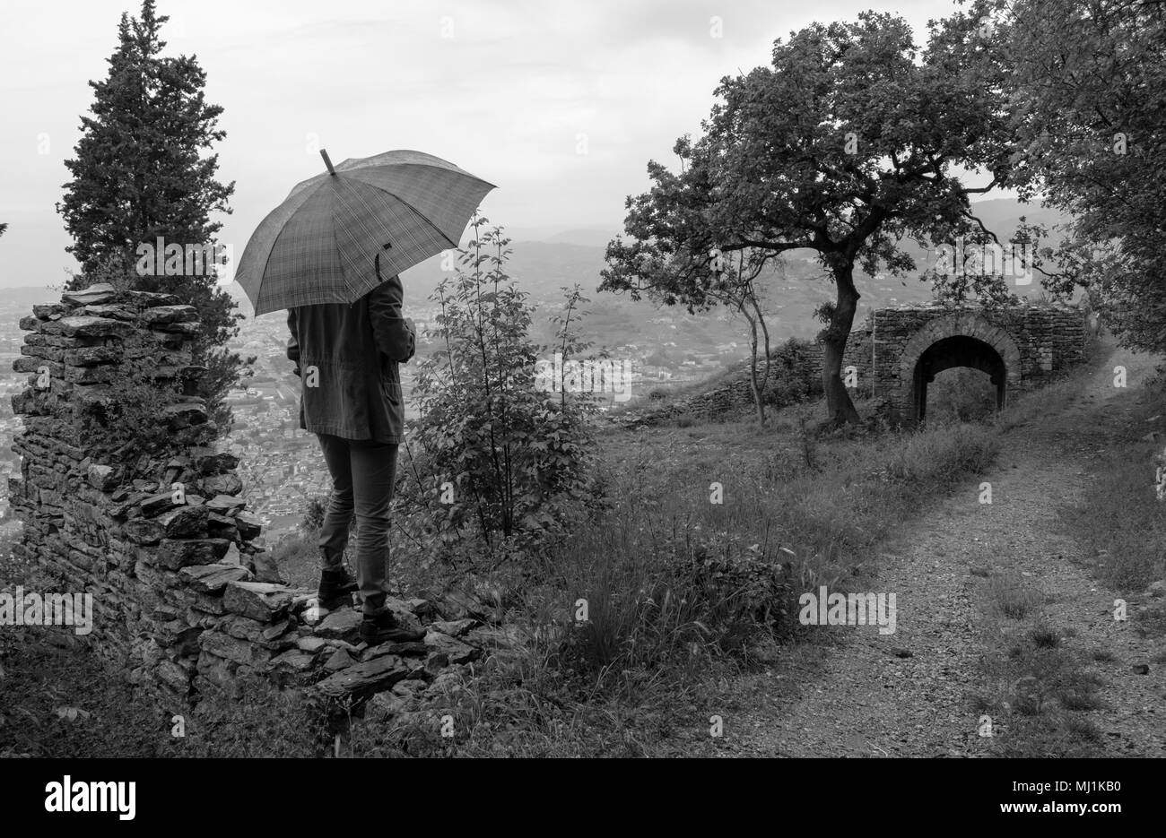 Un uomo solitario con un ombrello ed un cappotto lungo su una brutta muro di pietra osserva la vista di un antico portico durante un giorno di pioggia Foto Stock