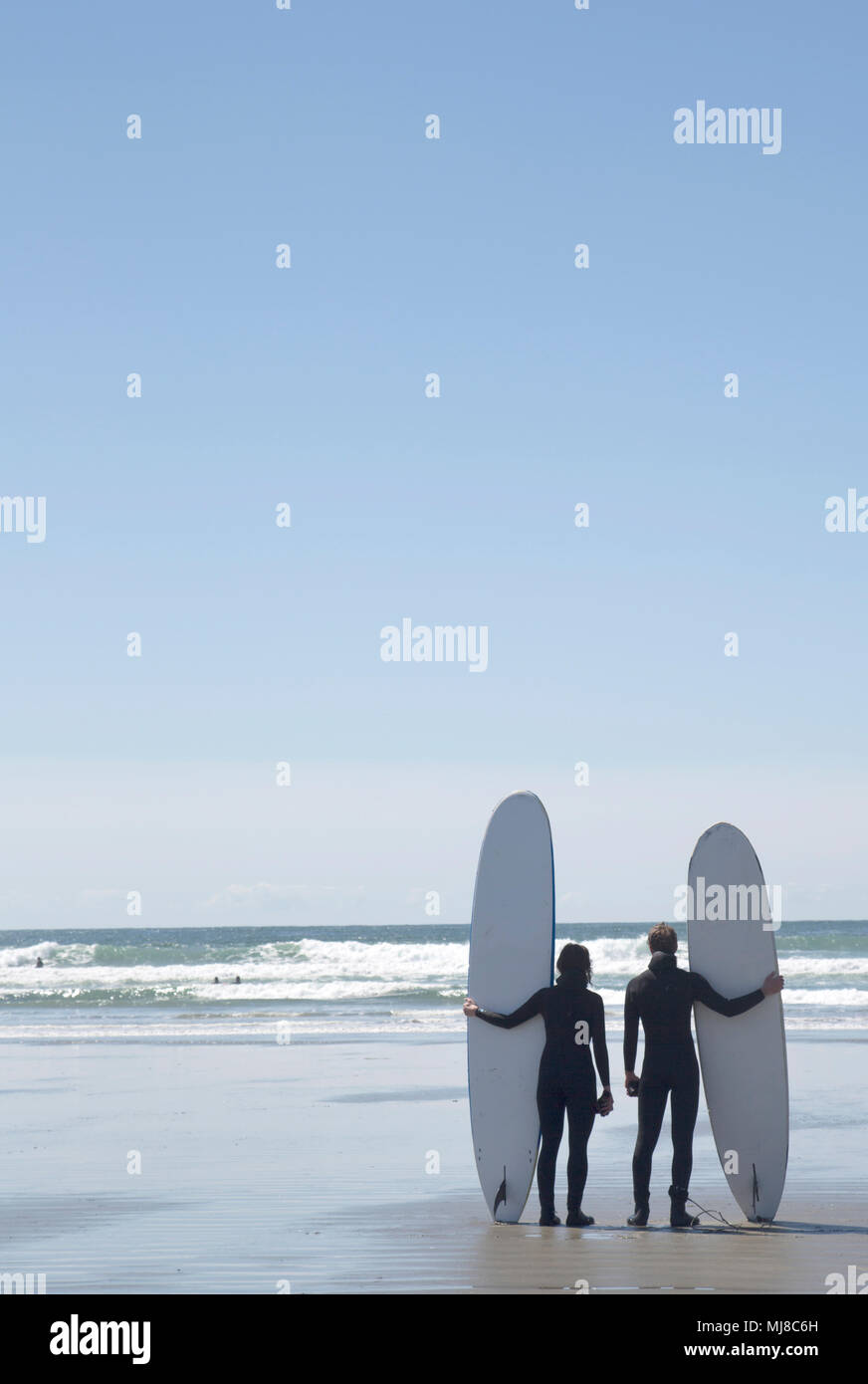Vista posteriore di un uomo e di una donna che indossa abiti bagnati in piedi a fianco a fianco sulla spiaggia sabbiosa dall'oceano, tenendo le tavole da surf. Foto Stock