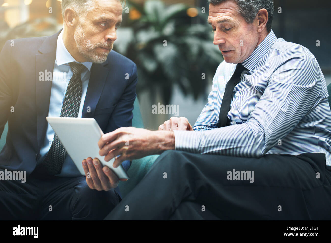 Due ben vestito imprenditori matura seduti su sedie in un ufficio lobby e discutere di lavorare insieme e utilizzando una tavoletta digitale Foto Stock