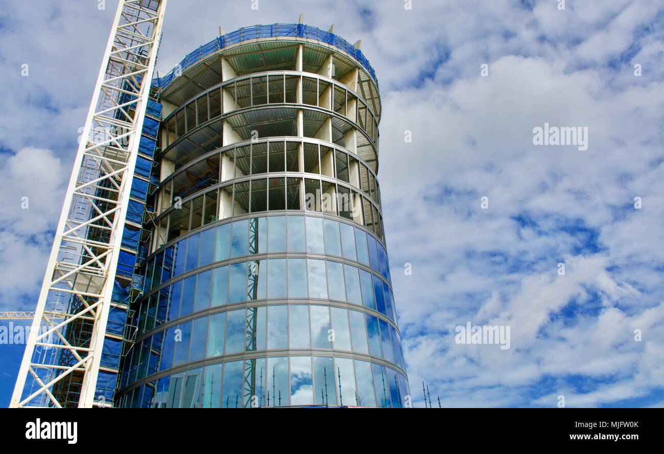 Costruzione di un nuovo cilindrico di vetro alto edificio con gru contro un nuvoloso cielo blu Foto Stock