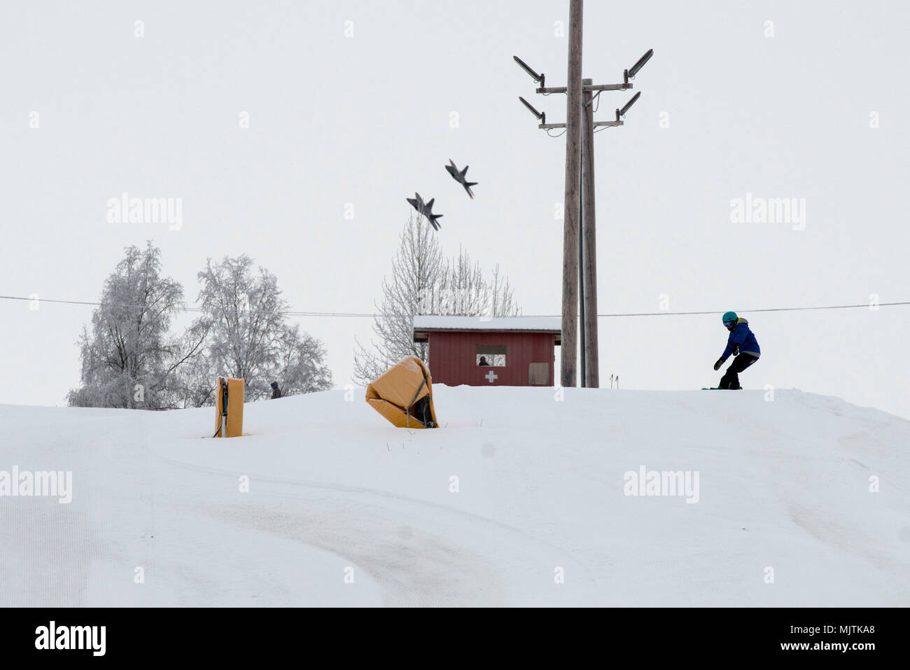 Uno snowboarder si prepara a andare giù Hillberg Area sciistica con F-22 rapaci in background durante il Winterfest a base comune Elmendorf-Richardson, Alaska, 21 dicembre 2017. Winterfest era un solstizio d'inverno celebrazione ospitato da Hillberg per chiunque con accesso di base e offerto una pletora di eventi da mezzogiorno al più tardi 8 p.m. (U.S. Air Force foto di Senior Airman Christopher R. Morales) Foto Stock