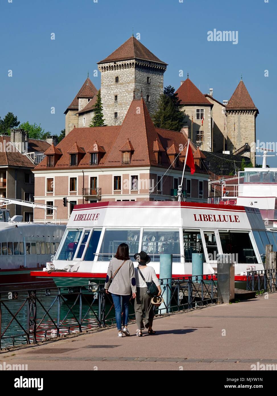 Due donne a piedi lungo la sponda del lago, passando per le imbarcazioni turistiche, con il castello di Annecy in aumento in background, Annecy, Francia Foto Stock