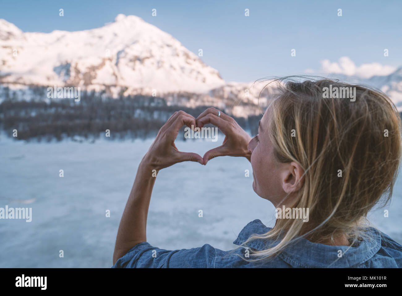 Giovane donna fare a forma di cuore dito telaio su lago ghiacciato e montagne dalle vette innevate. La gente ama la natura il concetto di ambiente. Girato in Svizzera Foto Stock