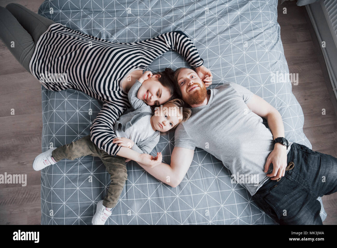 Vista superiore della giovane e bella madre, padre e la loro figlia guardando la fotocamera e sorridenti mentre giaceva sul letto testa a testa Foto Stock