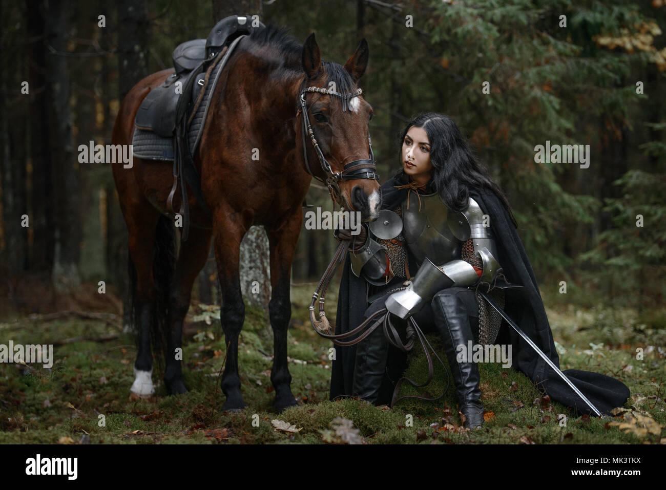 Una bella ragazza del guerriero con una spada che indossa chainmail e armatura con un cavallo in una foresta misteriosa Foto Stock