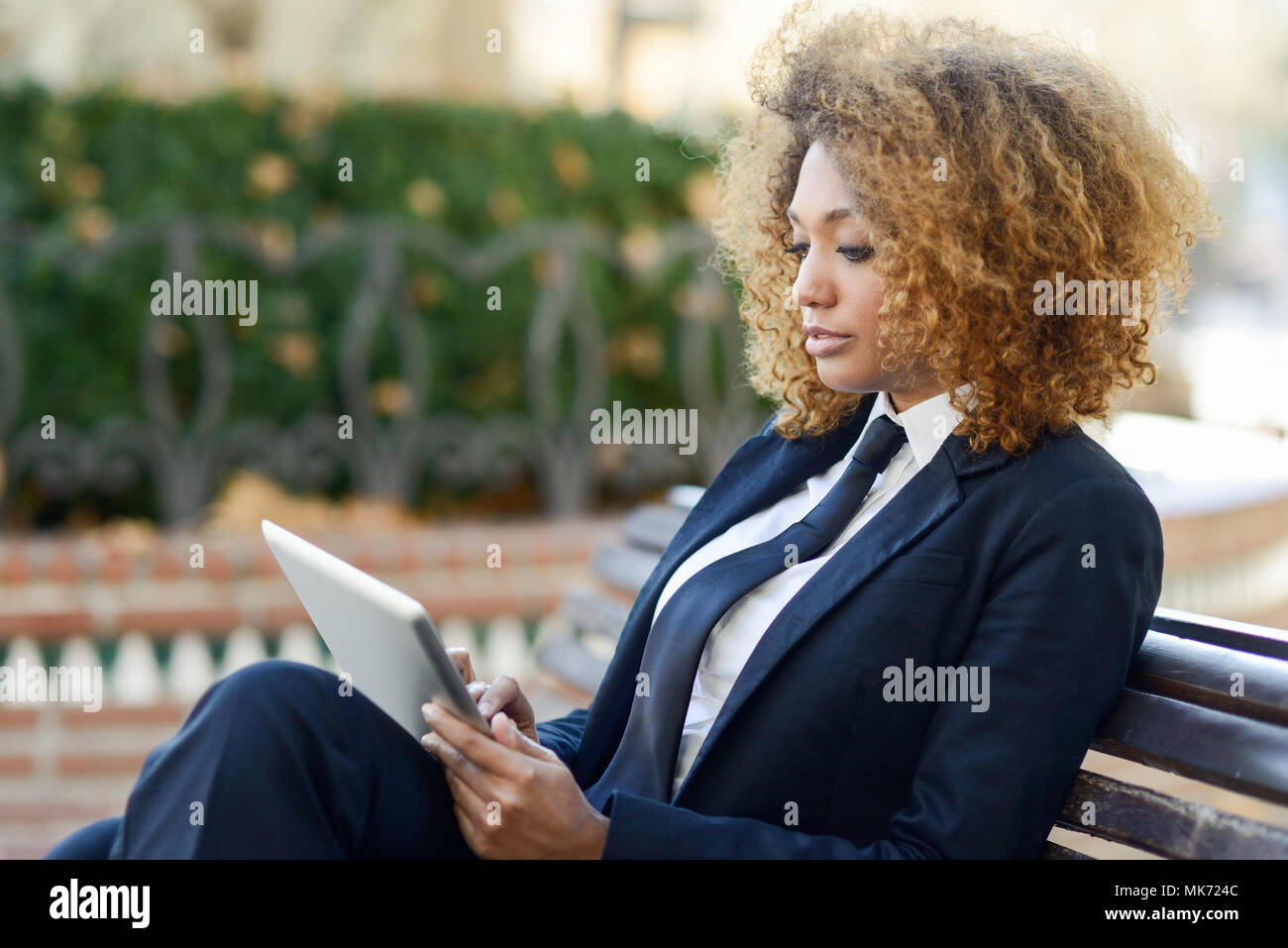Bella black capelli ricci donna africana utilizzando computer tavoletta su un banco di URBAN. Imprenditrice indossare tuta con pantaloni e cravatta, acconciatura afro. Foto Stock
