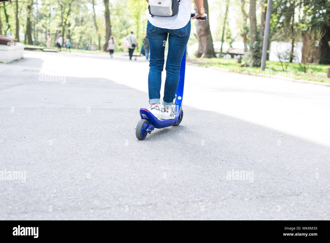 Persona che utilizza lo scooter elettrico e un smartwatch nel parco e sulla strada in una giornata di sole, con effetto bokeh di fondo. Foto Stock