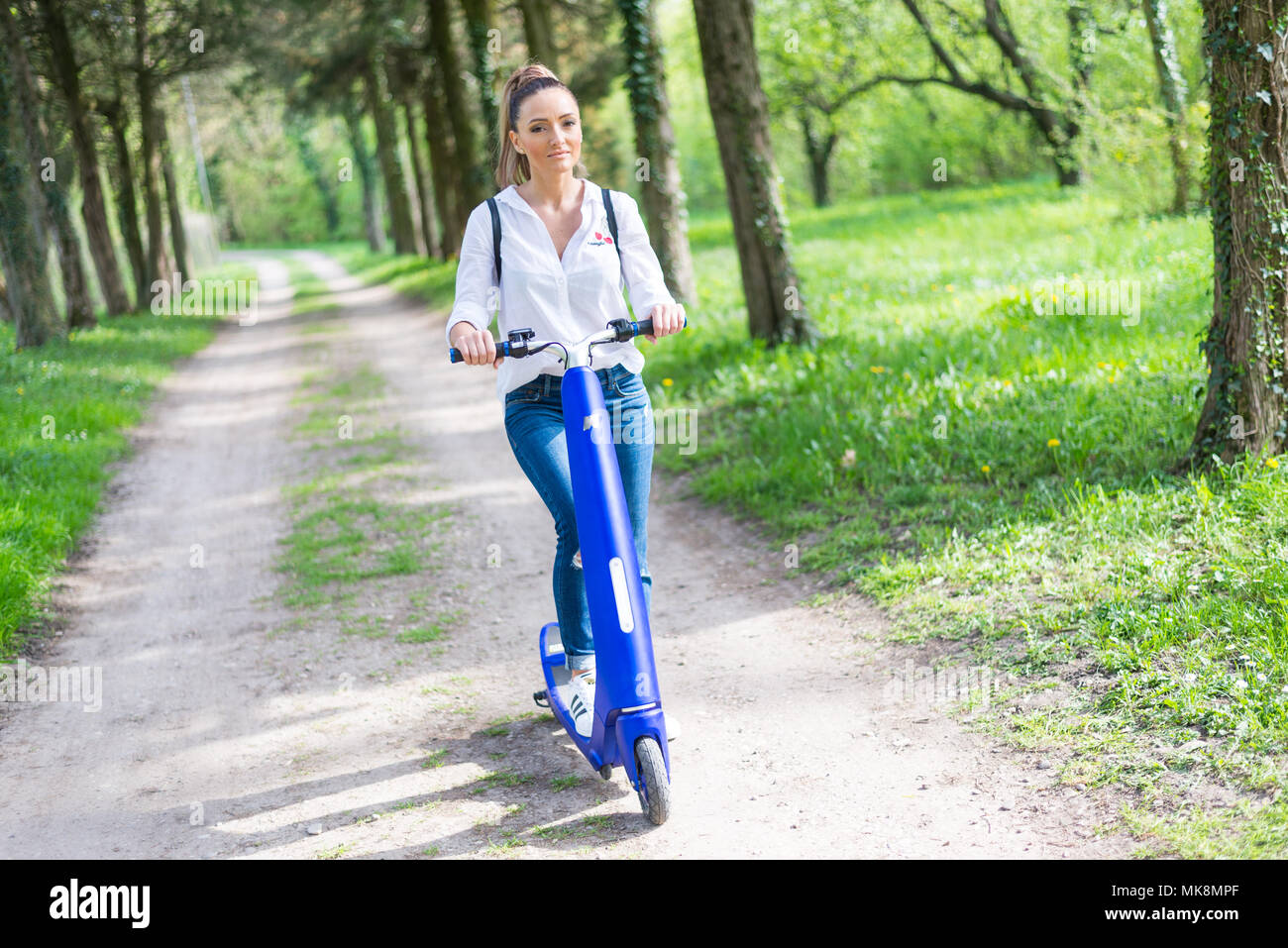 Persona che utilizza lo scooter elettrico e un smartwatch nel parco e sulla strada in una giornata di sole, con effetto bokeh di fondo. Foto Stock