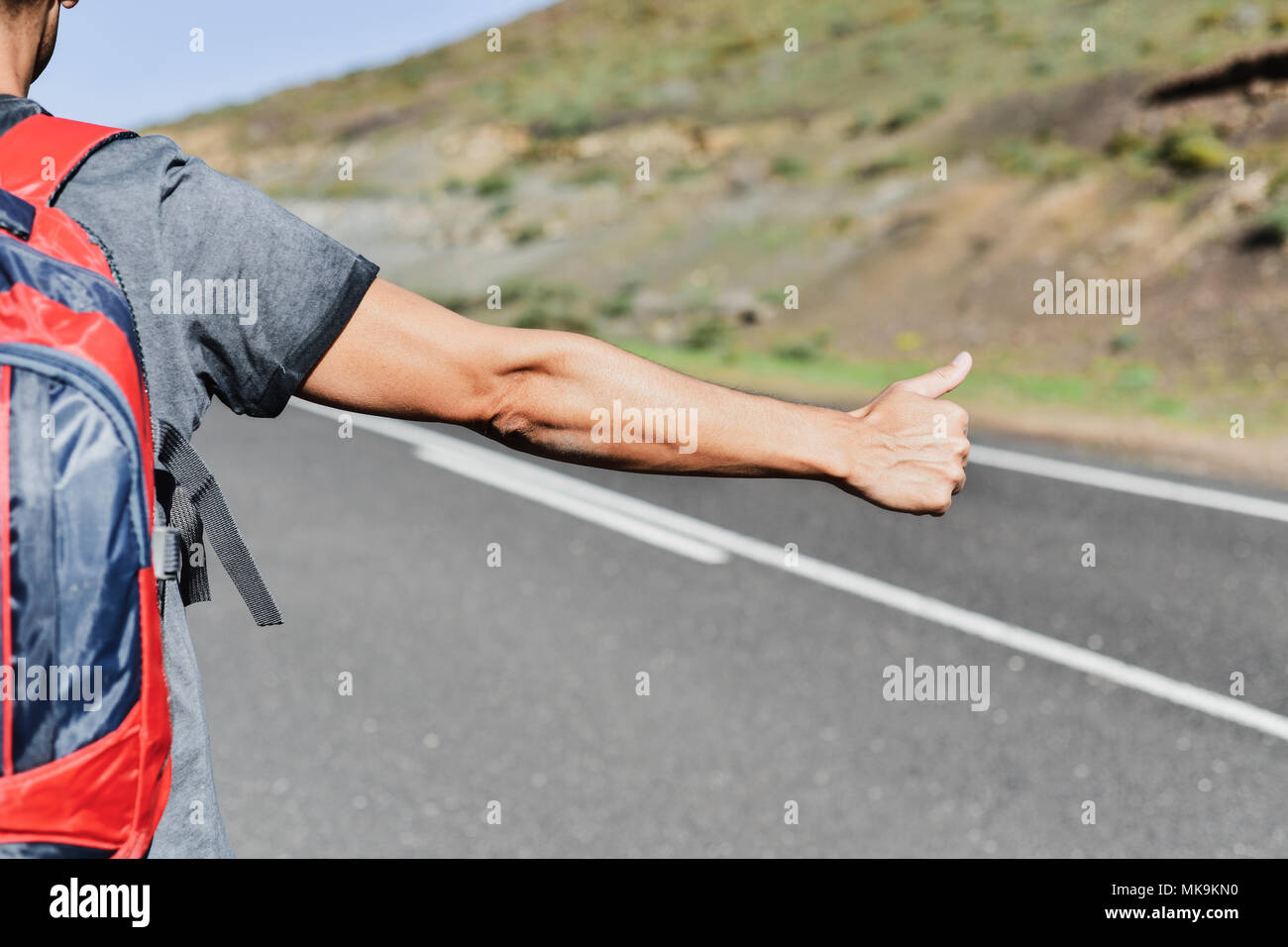 Primo piano di un giovane uomo caucasico, visto da dietro, portando uno zaino autostop in una strada secondaria, con il suo pollice in alto Foto Stock