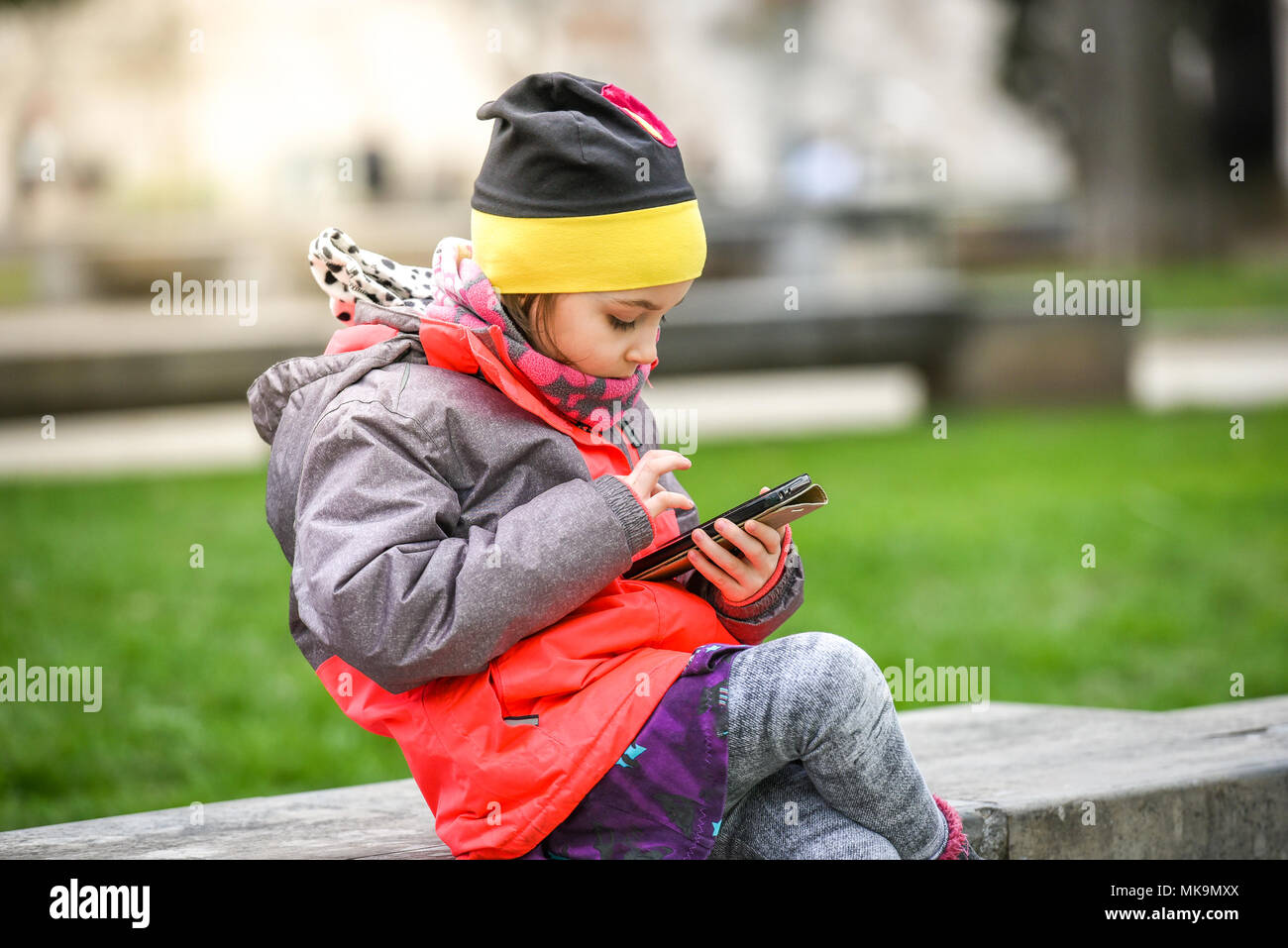 Bambina bambino usando un telefono cellulare nel parco pubblico. Invece di giocare al di fuori, il bambino è la riproduzione di giochi sul telefono, seduta nel banco in granito. Chil Foto Stock