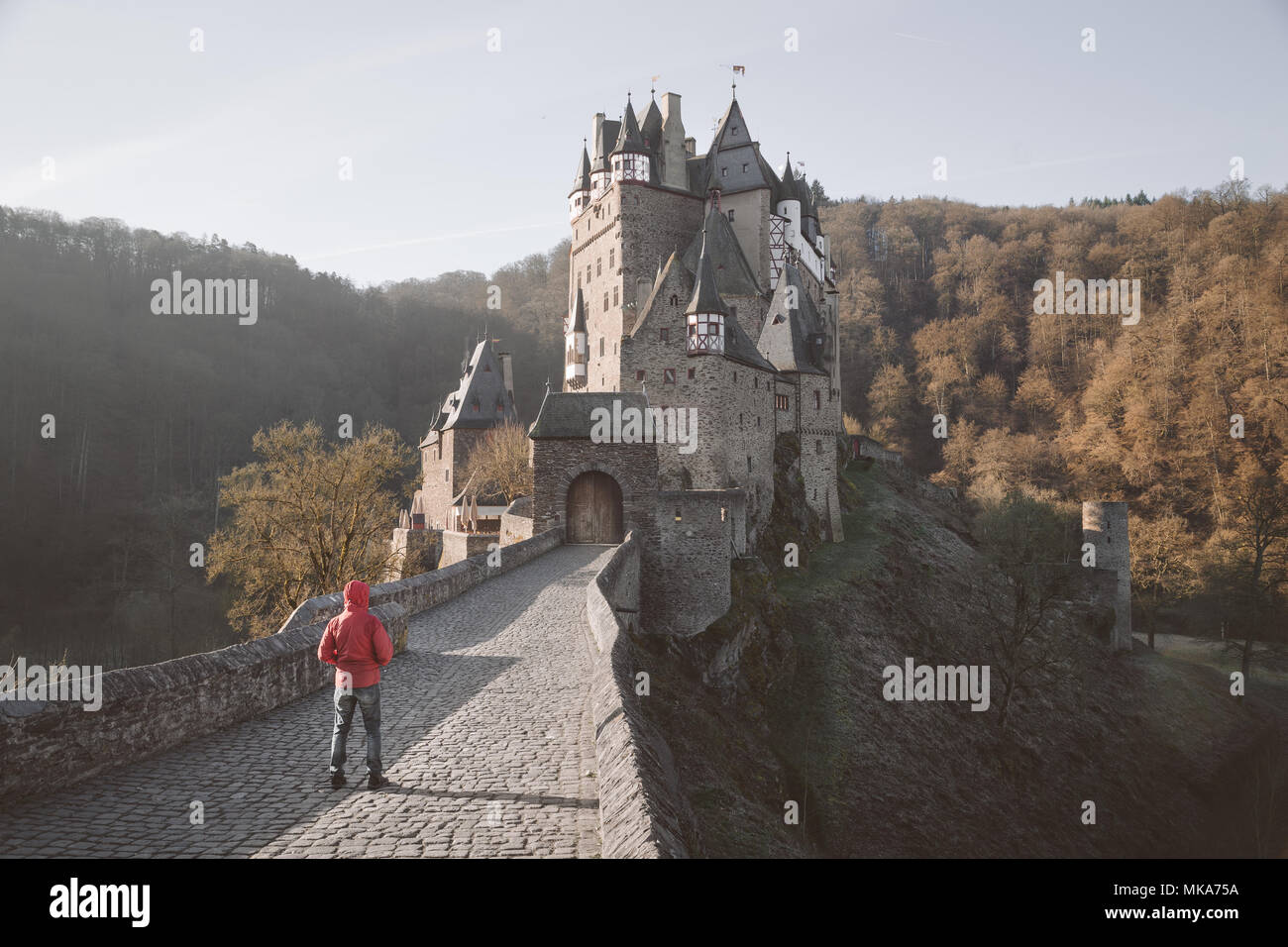 Visualizzazione classica del giovane in giacca rossa in piedi nella parte anteriore del famoso Castello Eltz a sunrise in caduta, Renania-Palatinato, Germania Foto Stock