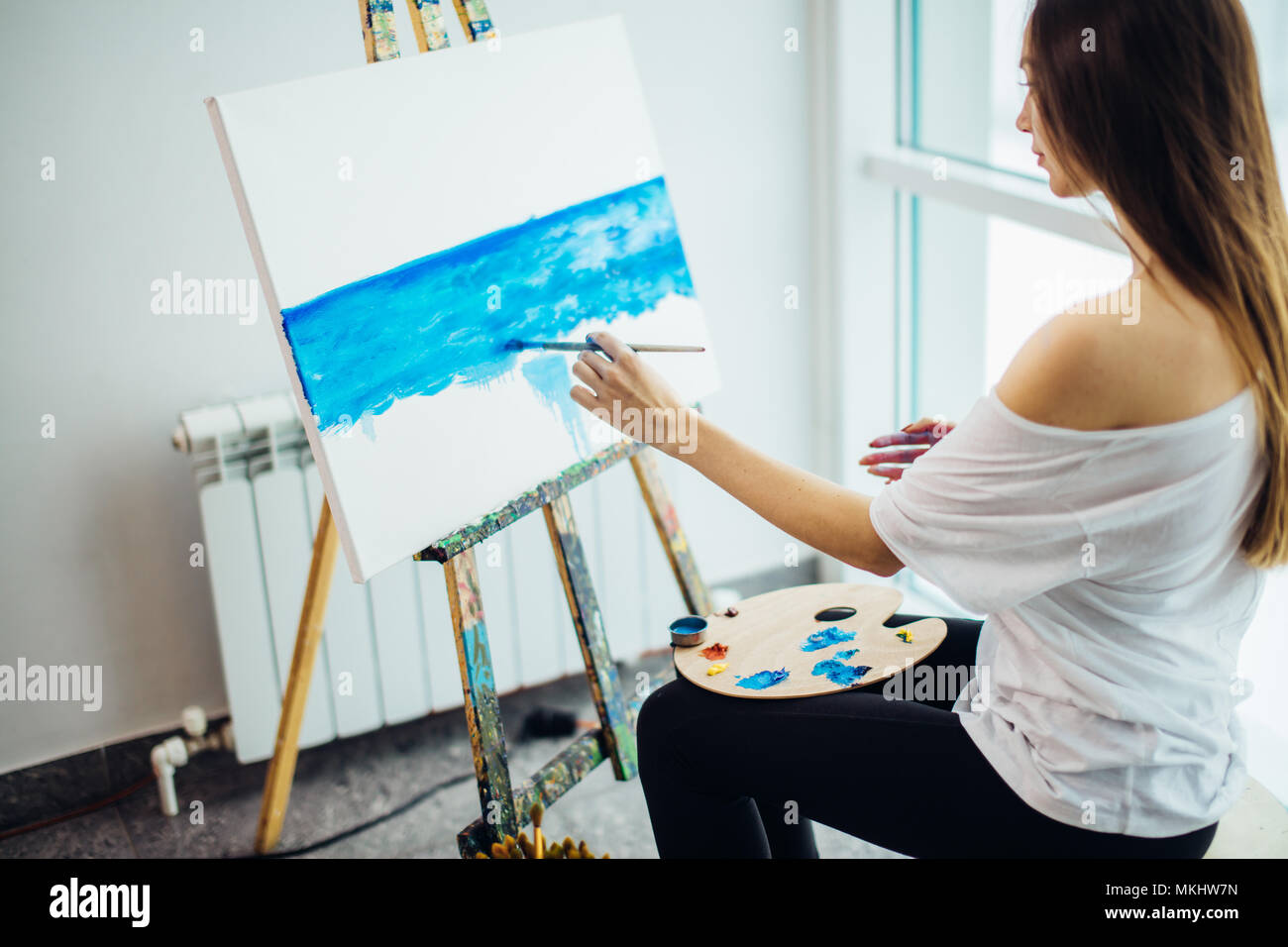 Giovane donna attraente artista pittura mare-scape su tela in officina, vista dal lato posteriore Foto Stock