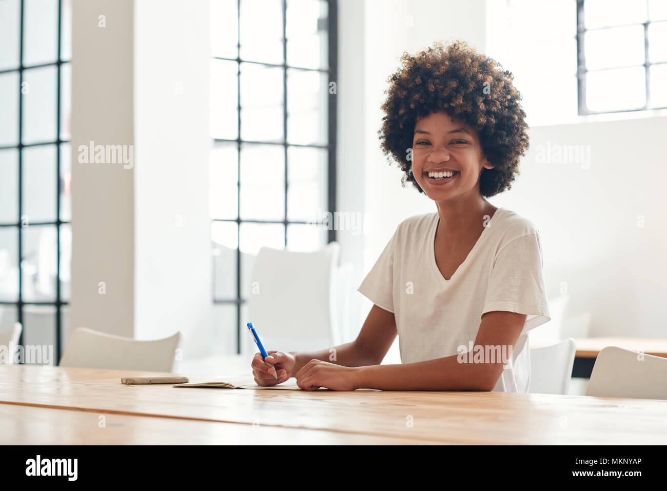 Ritratto di un sorridente giovane africana studentessa con un afro iscritto in un notebook mentre è seduto da solo al tavolo del campus Foto Stock