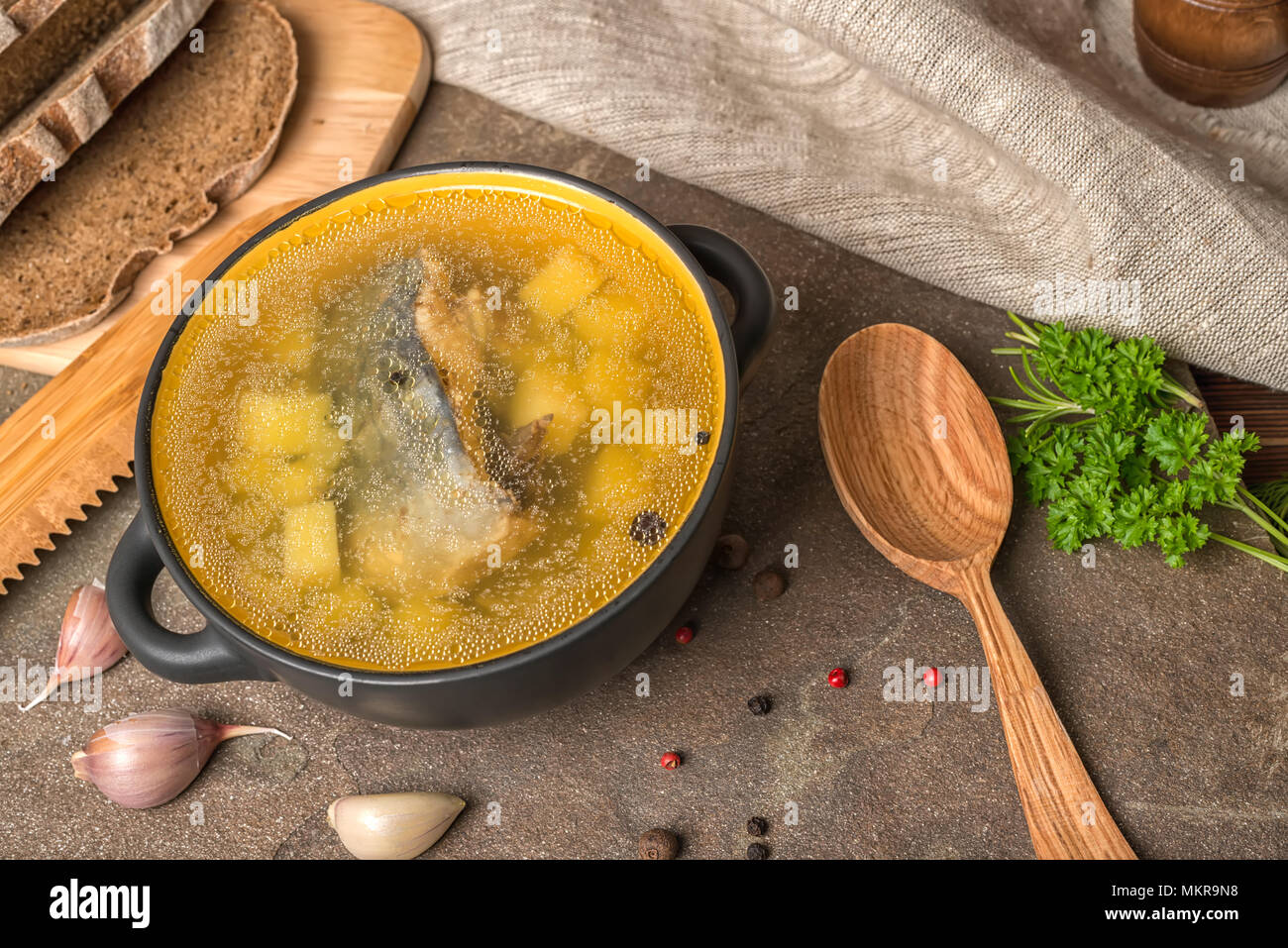 Trasparente la zuppa di pesce con lo storione, patate in piastra nera, decorato di aglio, prezzemolo in foglie, il cucchiaio di legno e tagliate il pane di segale, cucina etnica Foto Stock