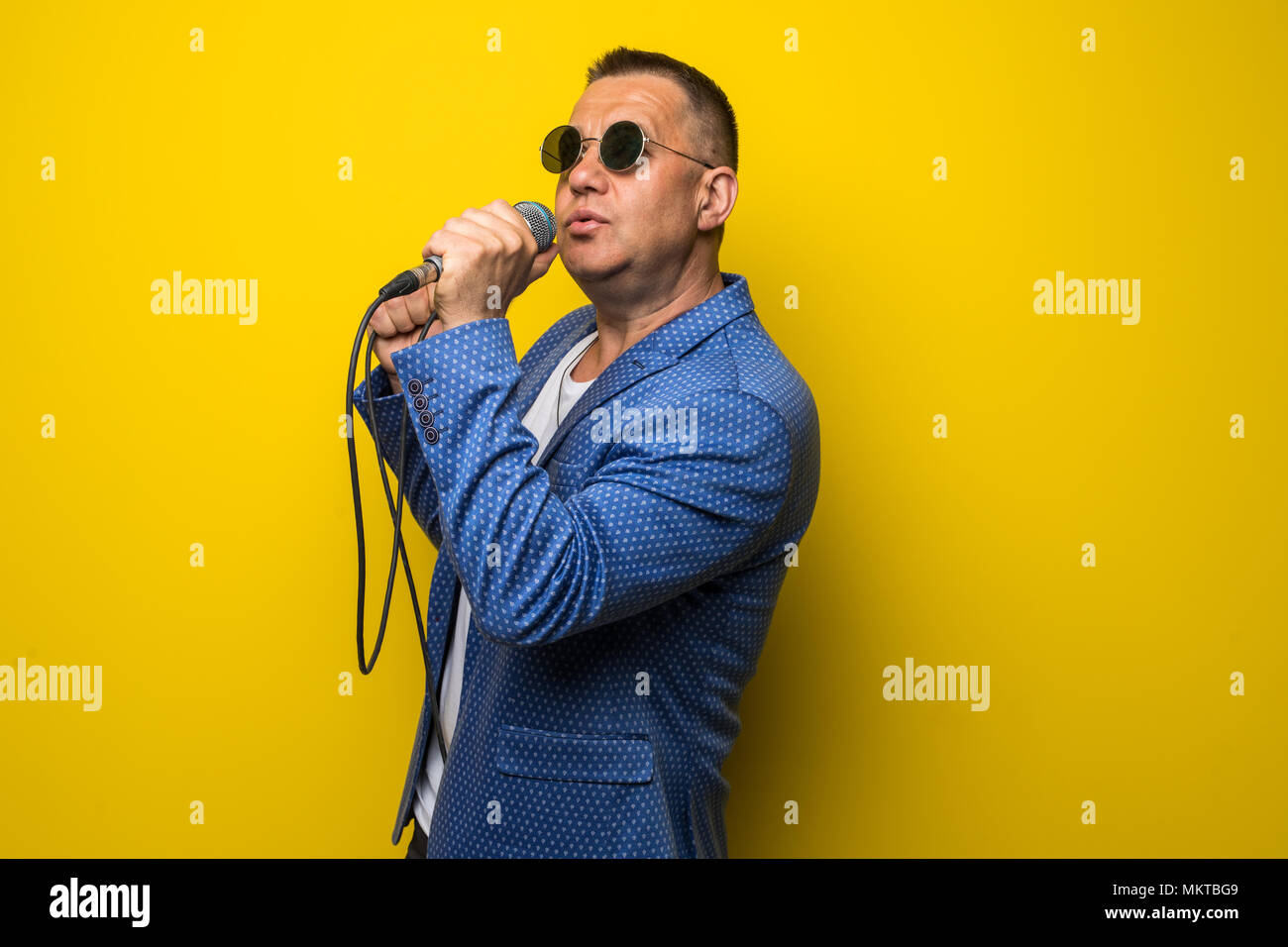 Portrai di matura età media uomo in tuta canti sul microfono isolato su sfondo giallo. Foto Stock