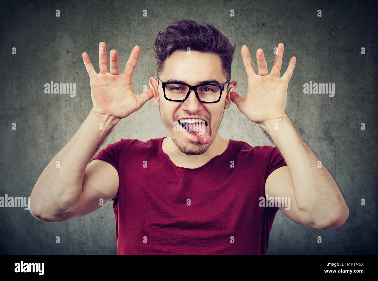 Ritratto di giovane uomo giocoso e smorfie che mostra la linguetta su sfondo grigio. Foto Stock