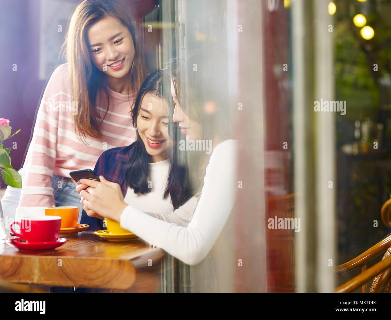Tre felice giovane e belle donne asiatiche seduta a tavola in chat parlando a giocare con il cellulare nel negozio di caffè o tè casa, shot attraverso la finestra glas Foto Stock