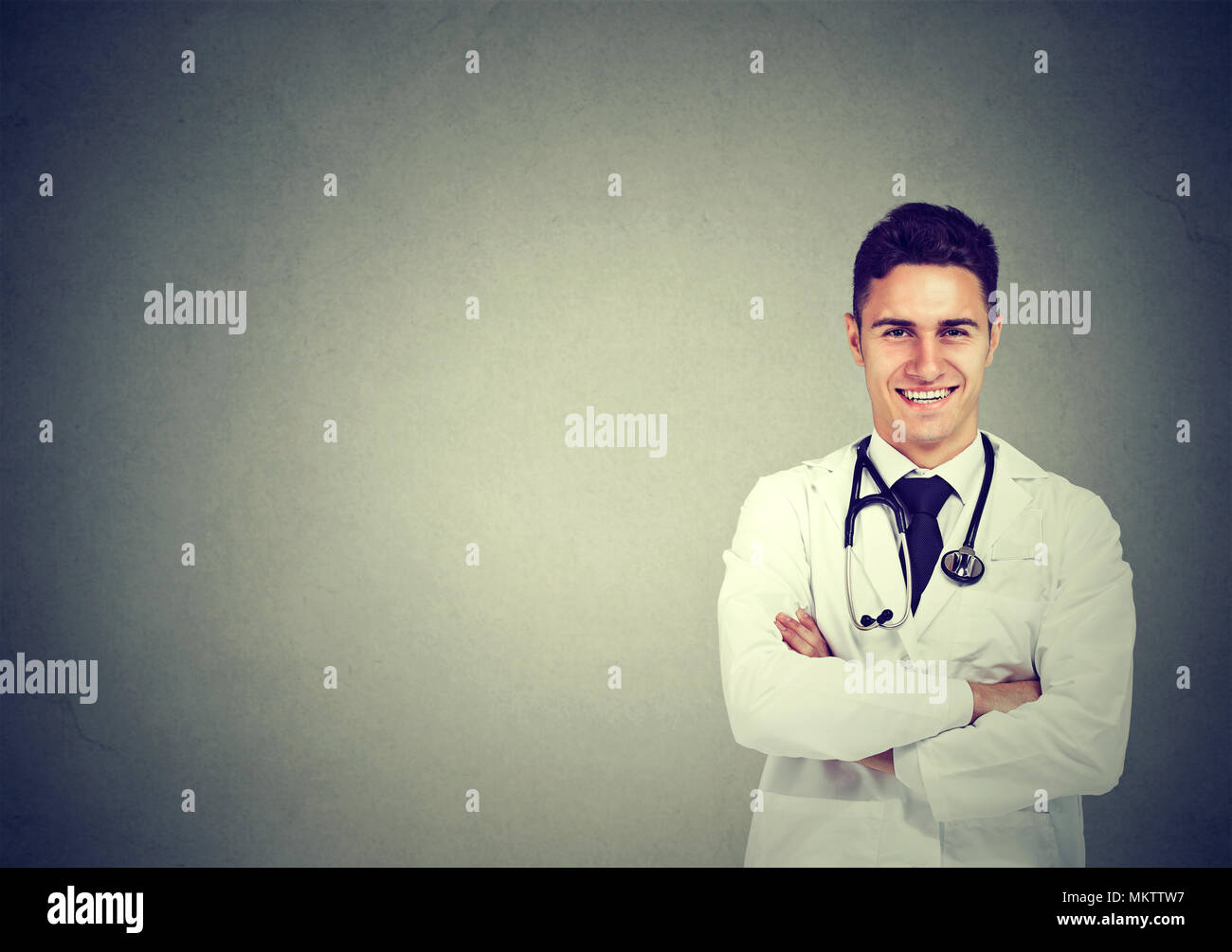 Medico sorridente in posa con le braccia incrociate sul muro grigio sfondo Foto Stock