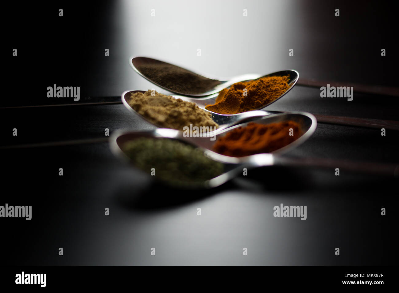 Spezie colorate in argento cucchiai, collocato su un tavoli in legno nero. Peperoncino, pepe, sale, basilico, senape Foto Stock