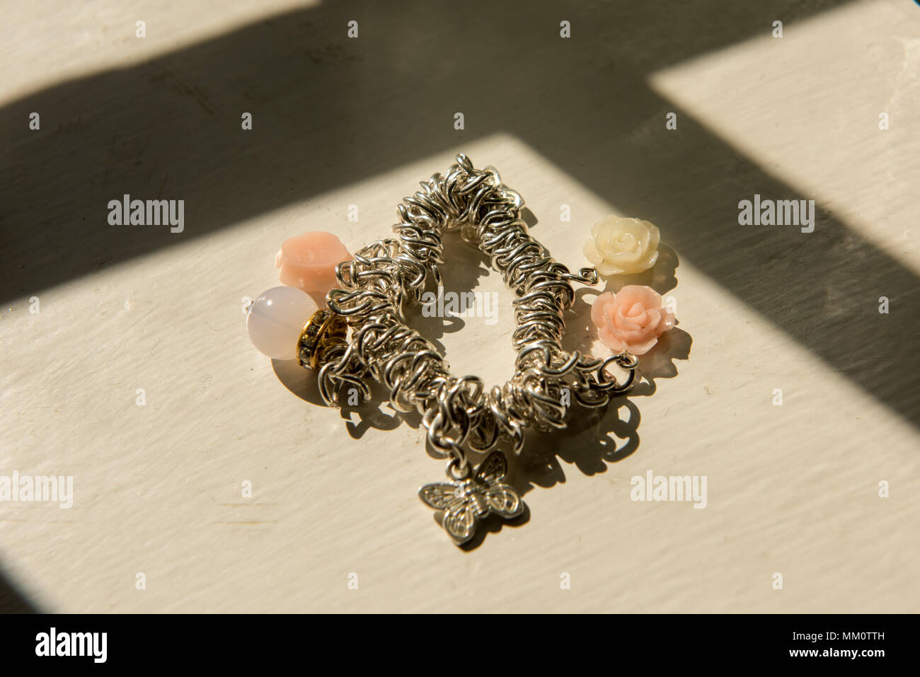 Bracciale ciondolo in argento con perline floreali e ciondolo a farfalla posizionato su un davanzale illuminato dal sole, che proietta ombre morbide Foto Stock