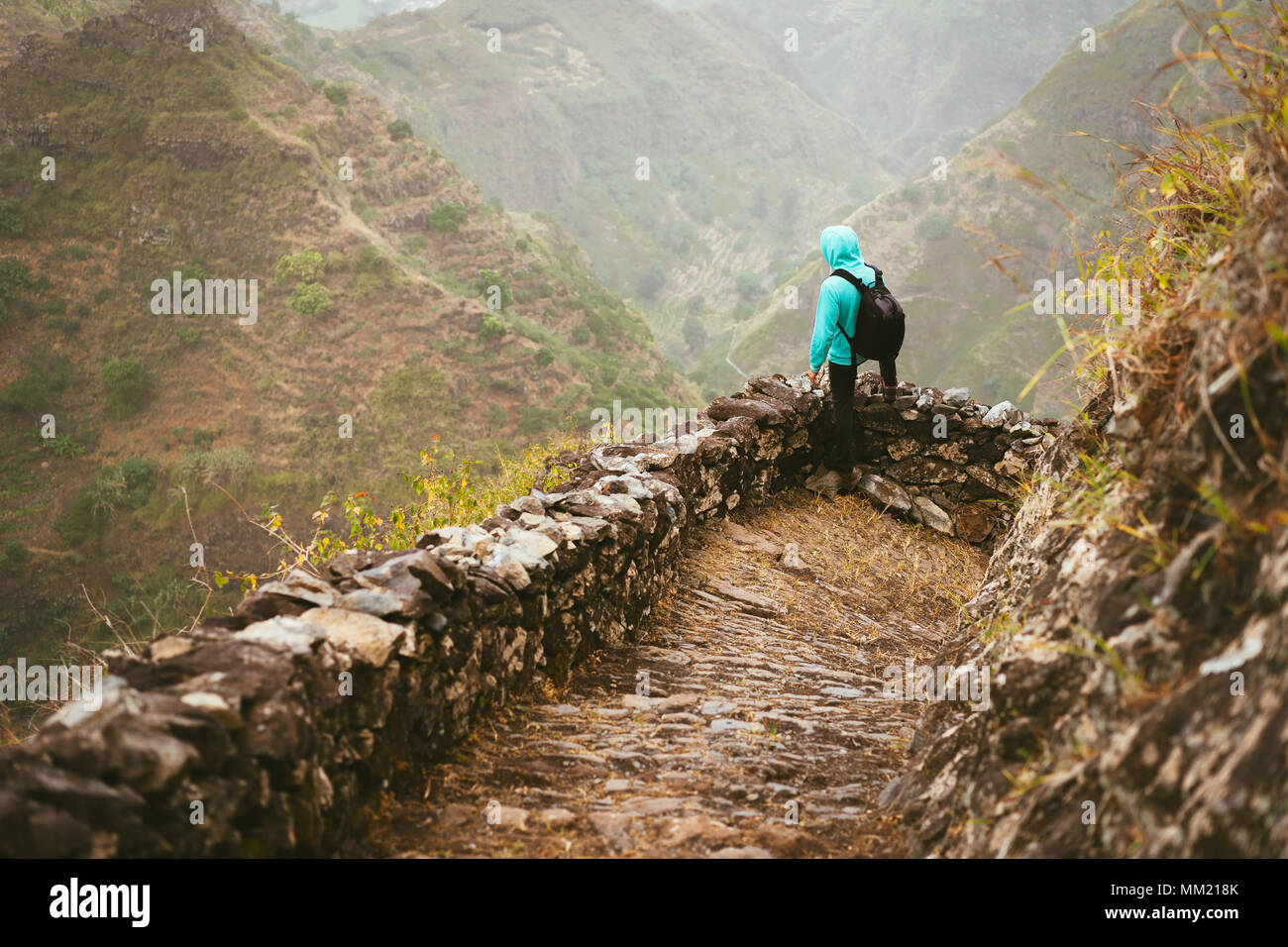 Escursionista con zaino sul bordo di montagna sentiero acciottolato guardando giù per la valle. Terreno roccioso di elevate catene montuose e profonde forre intorno a lui. Santo Antao Capo Verde Foto Stock