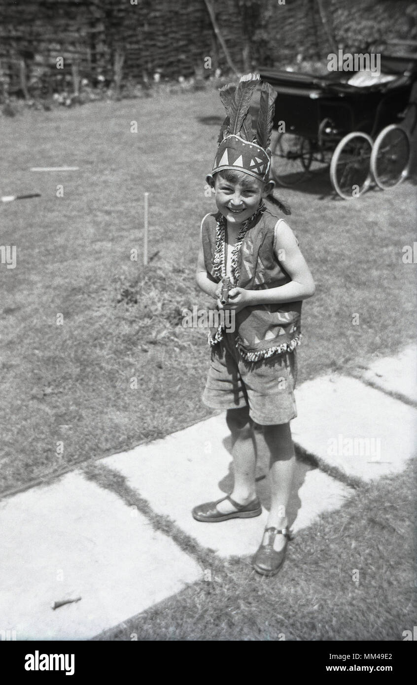 Degli anni Cinquanta, storico, giovane ragazzo in pantaloncini corti e indossa sandali fuori in un giardino vestito come un indiano con un gilè e piuma hat, Inghilterra. Regno Unito. Un bambino della PRAM è seduta sul prato in background. Foto Stock