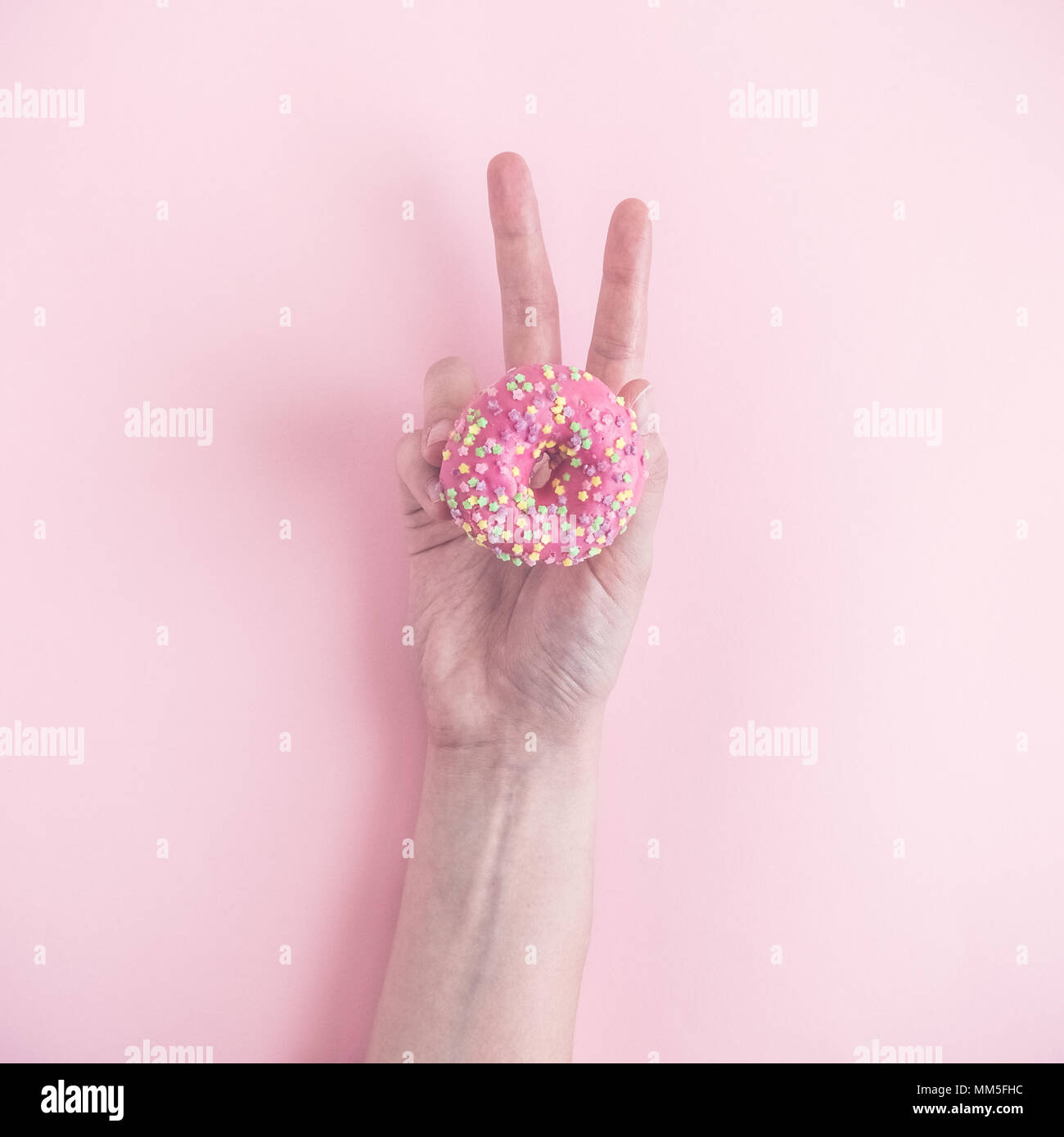 Stretta di mano femminile azienda piccole ciambelle e mostrando segno di vittoria con le dita sul rosa pastello minima di sfondo concetto creativo. Foto Stock