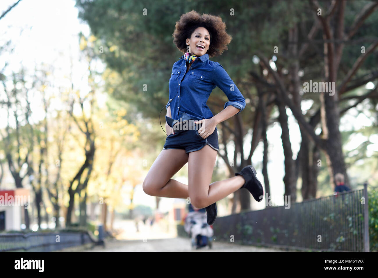 Giovane donna nera con acconciatura afro jumping in background urbano. Miscelati donna che indossa maglietta blu e pantaloncini. Porta femmina cuffie divertenti. Foto Stock