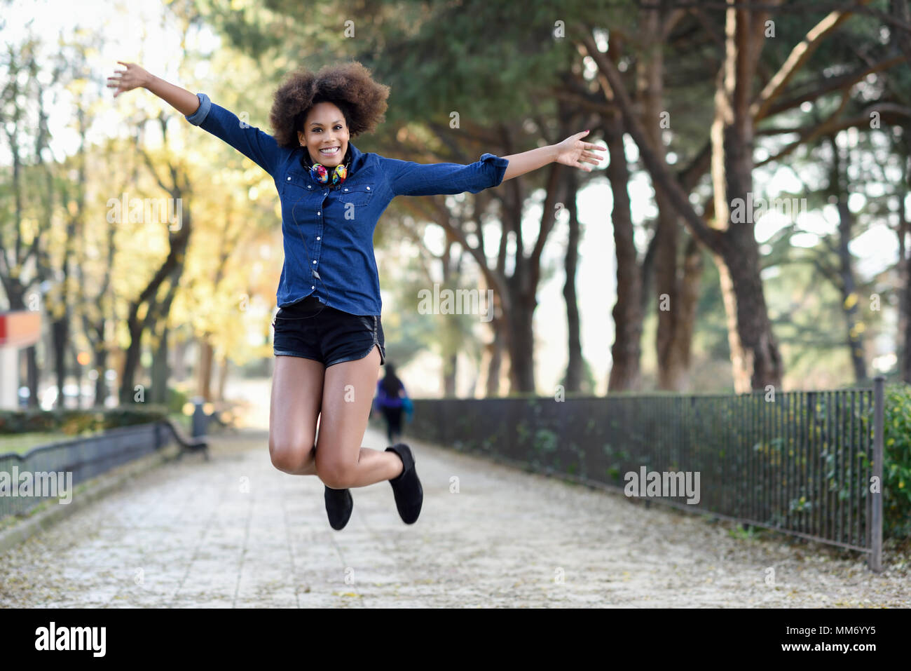 Giovane donna nera con acconciatura afro jumping in background urbano con le braccia aperte. Miscelati donna che indossa maglietta blu e pantaloncini. Porta femmina funny hea Foto Stock