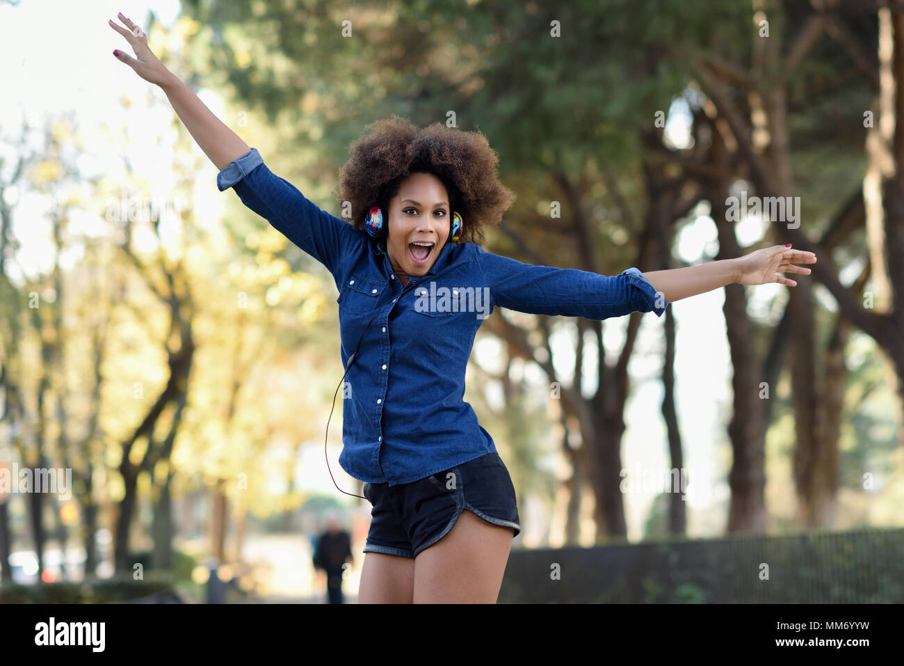 Giovane donna nera con acconciatura afro jumping in background urbano con le braccia aperte. Miscelati donna che indossa maglietta blu e pantaloncini. Porta femmina funny hea Foto Stock