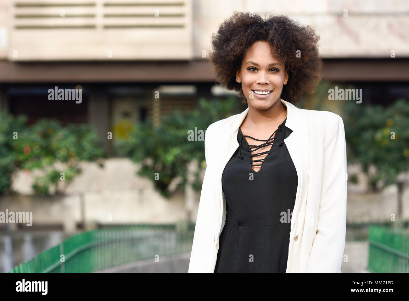 Giovane donna nera con acconciatura afro sorridente in background urbano.  Ragazza misti indossando giacca bianca e abito nero Foto stock - Alamy
