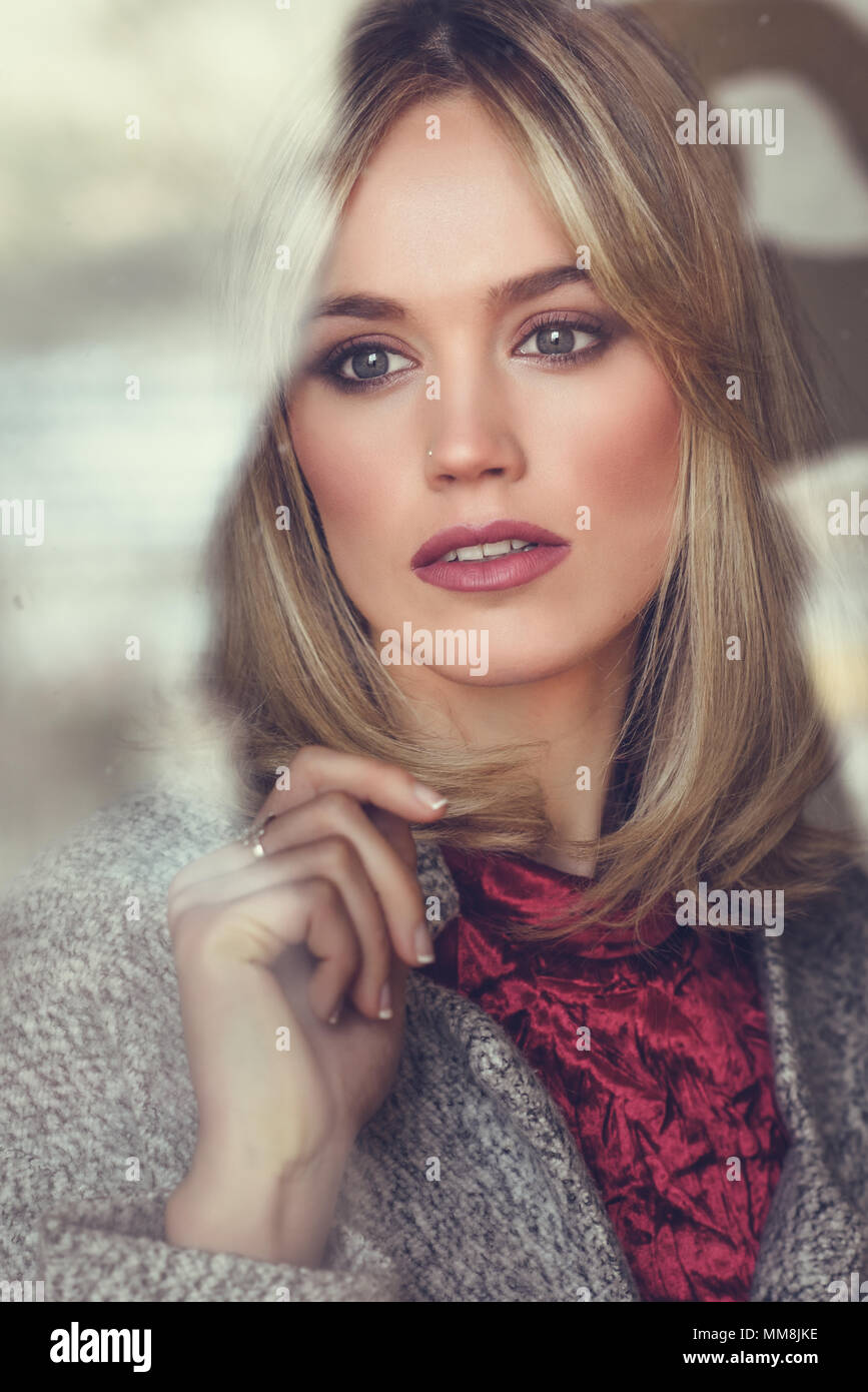 Considerato giovane donna bionda con gli occhi persi dalla finestra. Bella ragazza a casa indossando abiti invernali. Bella femmina con i capelli dritti e blu Foto Stock