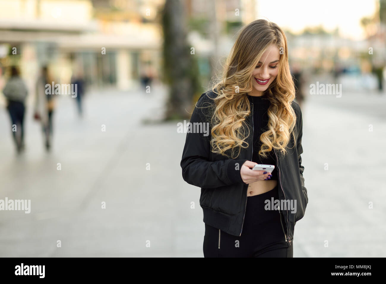 Donna bionda texting con il suo smart phone in background urbano. Bellissima fanciulla indossando giacca nera a piedi in strada. Pretty donne russe Foto Stock