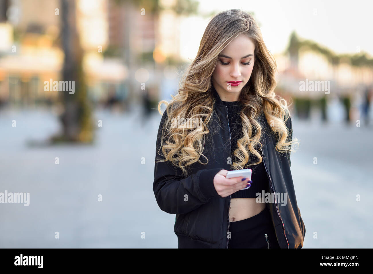 Donna bionda texting con il suo smart phone in background urbano. Bellissima fanciulla indossando giacca nera a piedi in strada. Pretty donne russe Foto Stock
