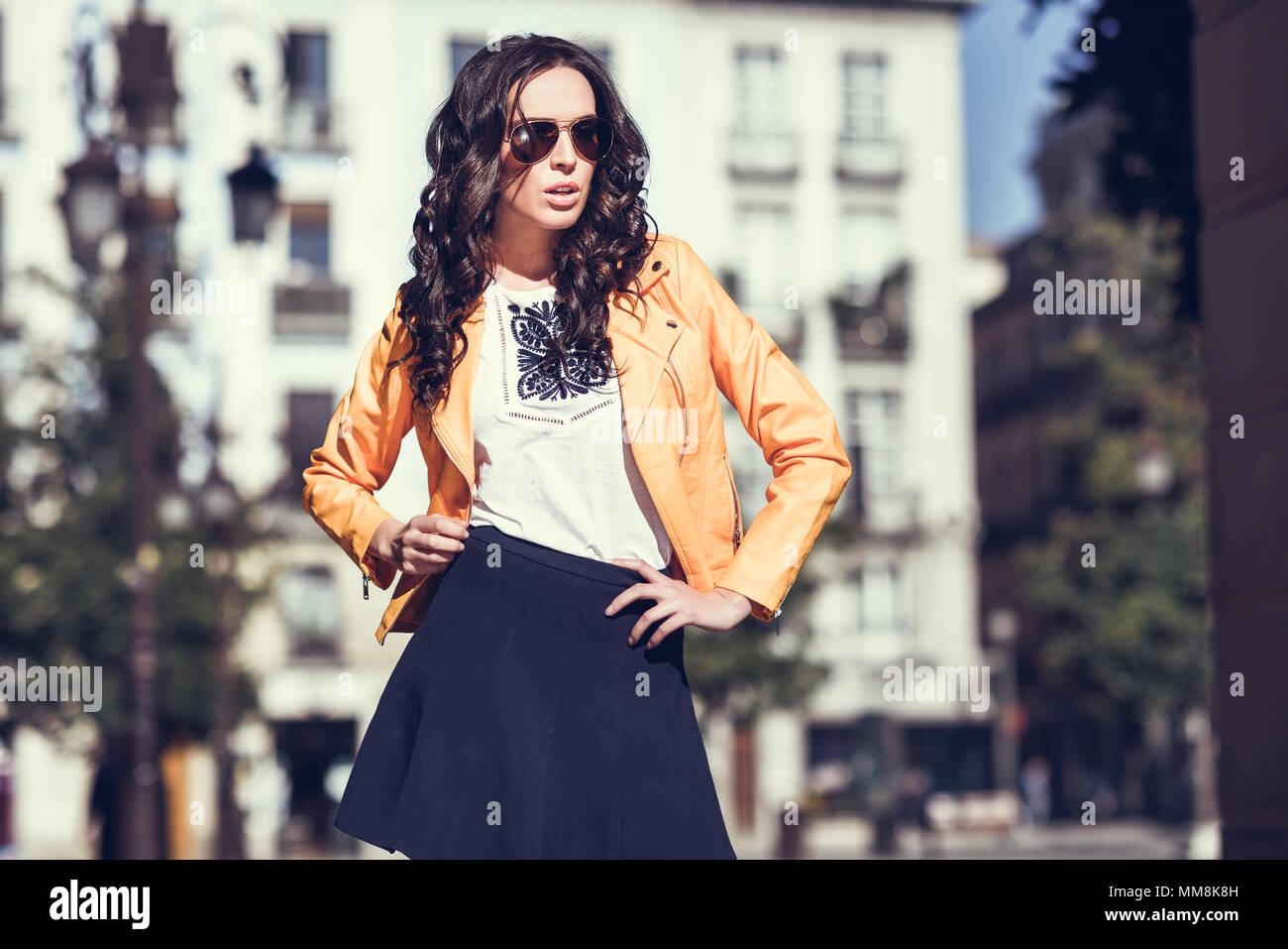 Bruna giovane donna con occhiali da sole. Ragazza, modello di moda, indossando orange Moderna giacca e gonna blu, stando in piedi in background urbano. Foto Stock