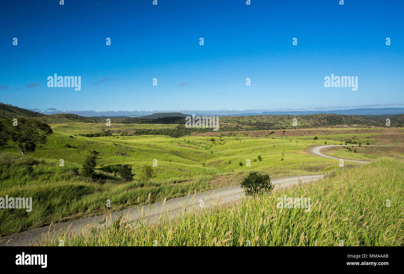 Vasto paesaggio con avvolgimento strada di ghiaia severing basse colline ammanta con erbe di smeraldo sotto il cielo blu nel Queensland del nord Foto Stock