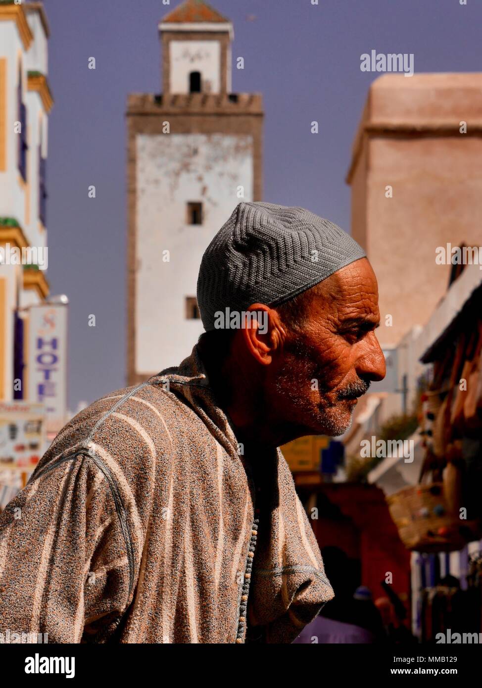 Primo piano di un uomo marocchino in un abito tradizionale passeggiando davanti alla torre della moschea a Essaouira, Marocco Foto Stock