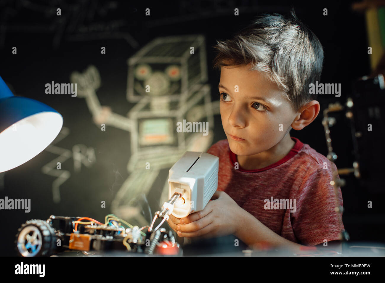 Riflessivo ragazzo lavorando sul suo progetto elettronico con una pistola per saldatura. Smart scolaro la saldatura di parti metalliche del suo giocattolo auto insieme a casa. Foto Stock