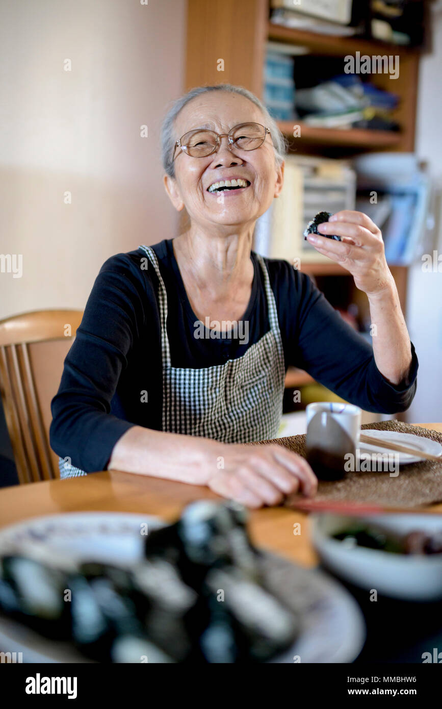 Donna anziana seduta al tavolo della cucina, mangiare sushi, sorridente alla fotocamera. Foto Stock
