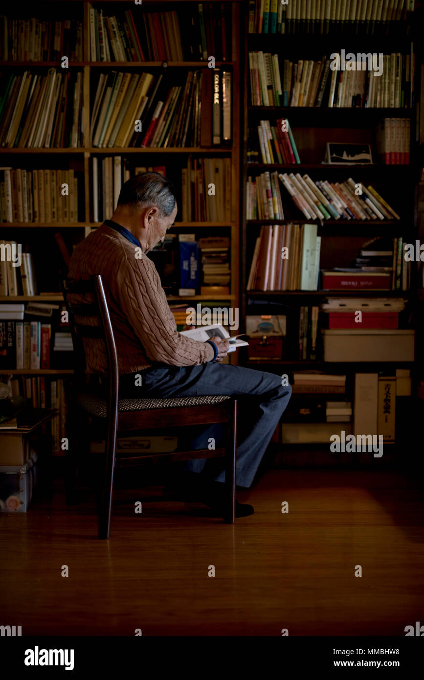 Anziani uomo seduto su una sedia nella parte anteriore della libreria, la lettura del libro. Foto Stock