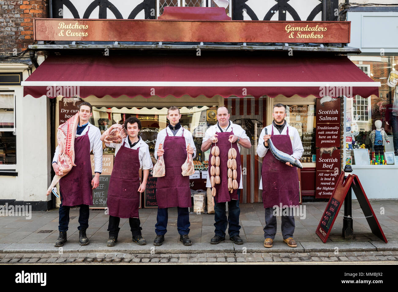 Gruppo di uomini, macellai e pescheria, indossando grembiuli, in piedi sul marciapiede al di fuori di una macelleria, tenendo i pezzi di carne e pesce, guardando a ca Foto Stock