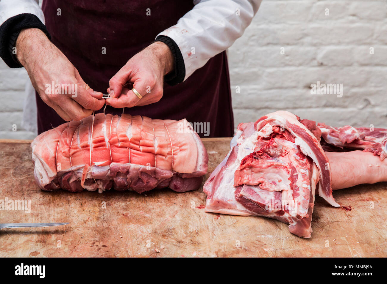 Chiusura del macellaio grembiule da indossare in piedi presso un macellaio in legno del blocco, preparare un laminato pancetta di maiale utilizzando la stringa. Foto Stock