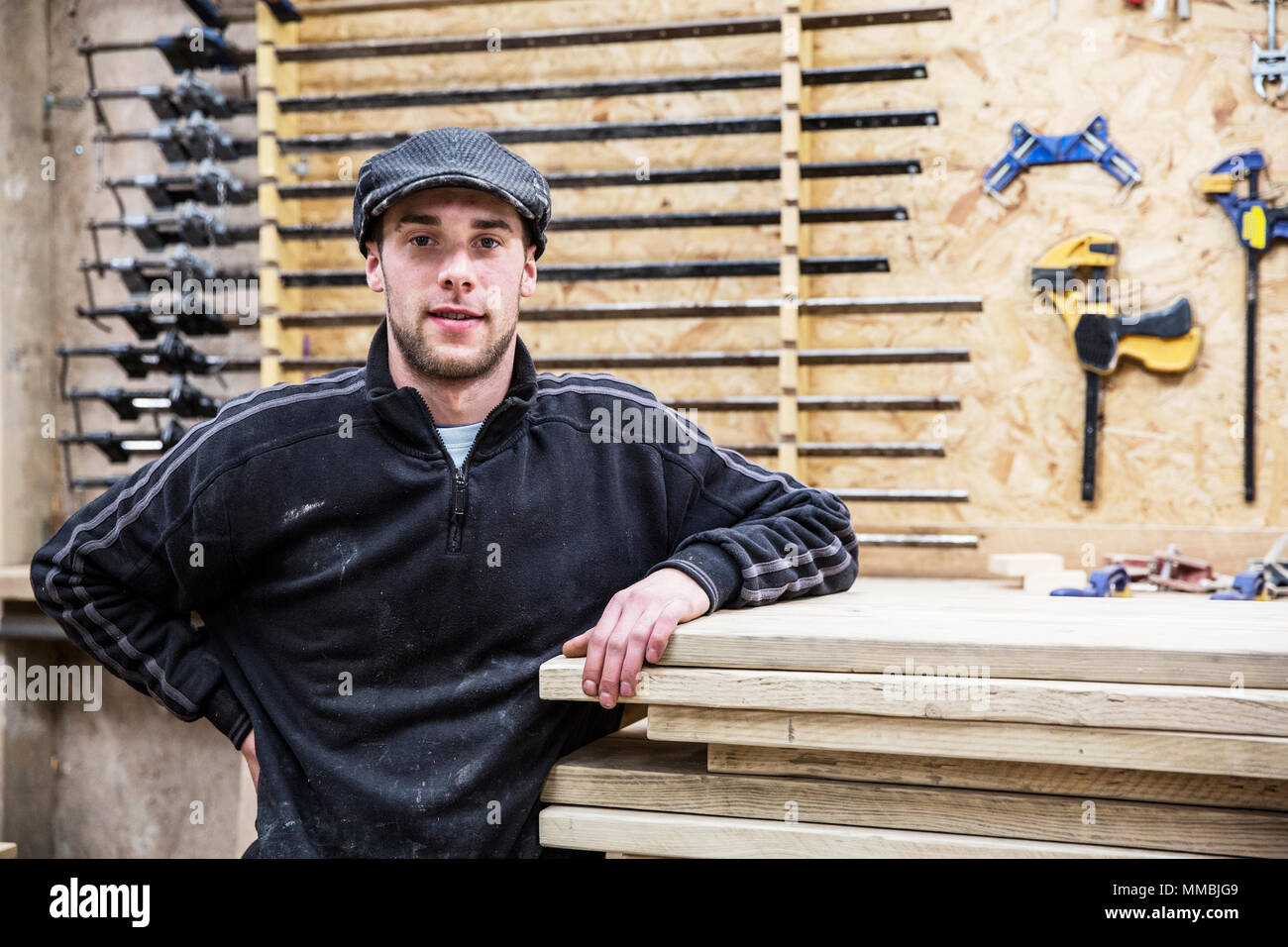 Uomo che indossa il tappo piatto in piedi accanto a una pila di pannelli in legno, guardando la telecamera, strumenti in background. Foto Stock